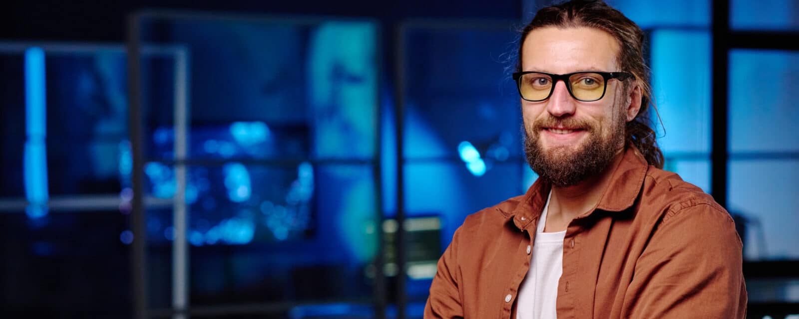 Homme avec lunettes noires et chemise marron souriant devant un fond bleu flou technologique.