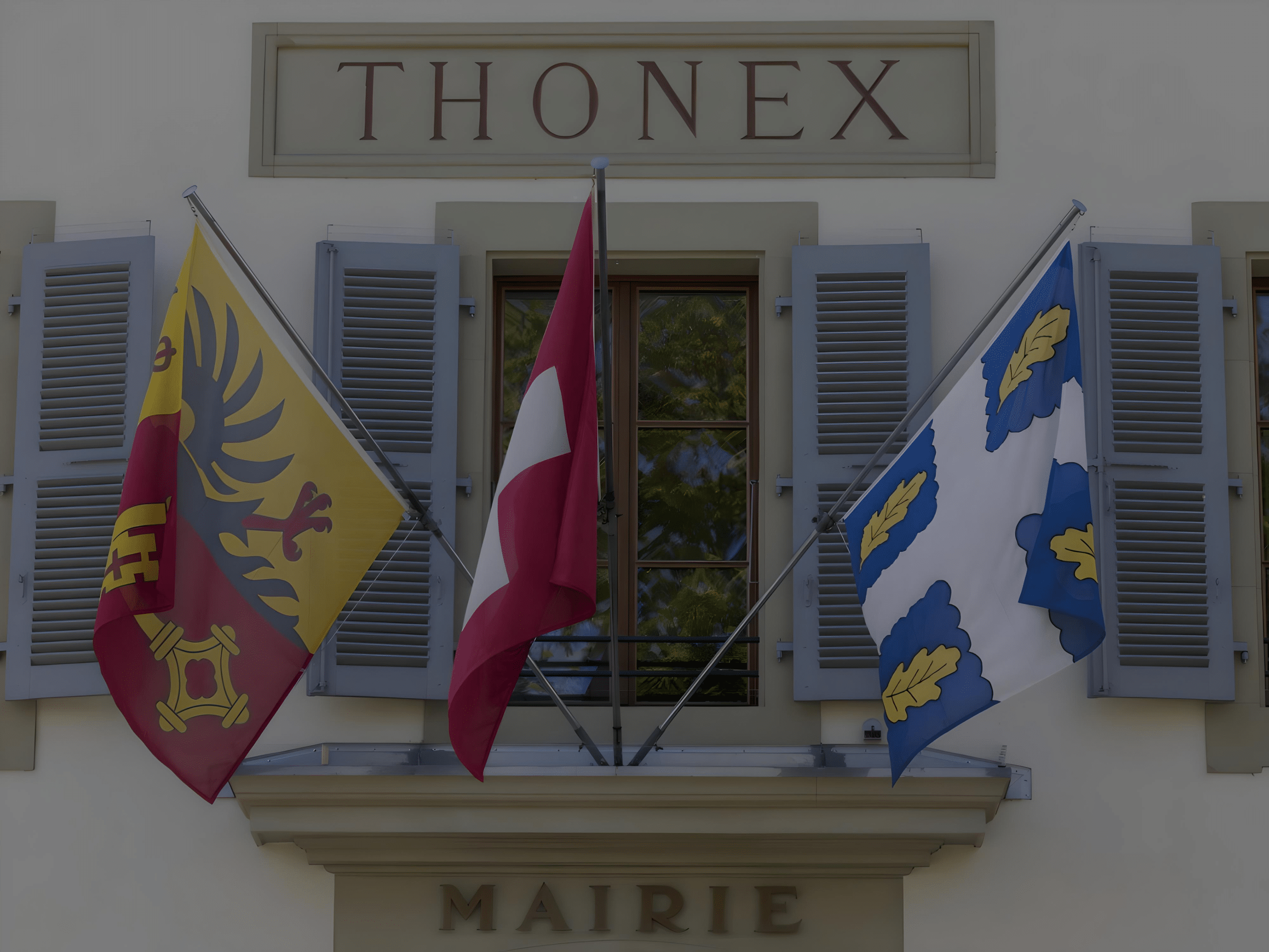 Façade de la mairie de Thonex avec trois drapeaux et volets gris autour d'une fenêtre.