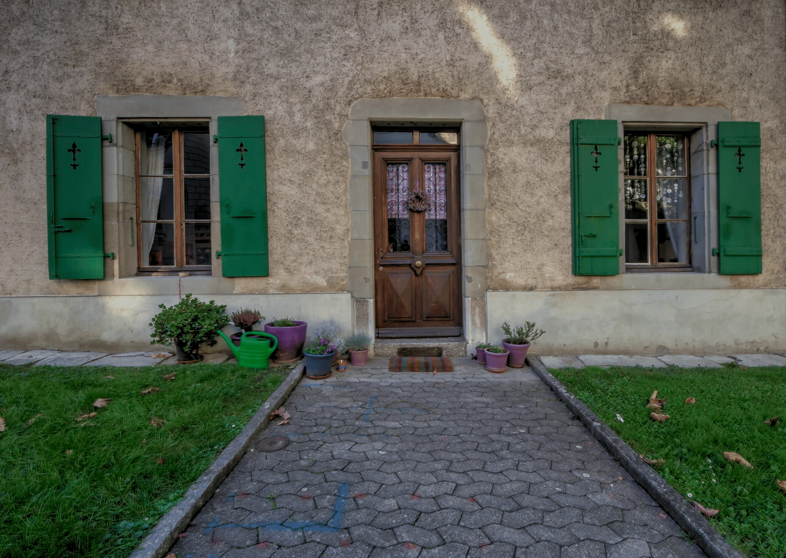 Façade de maison avec porte en bois, fenêtres à volets verts et pots de fleurs.