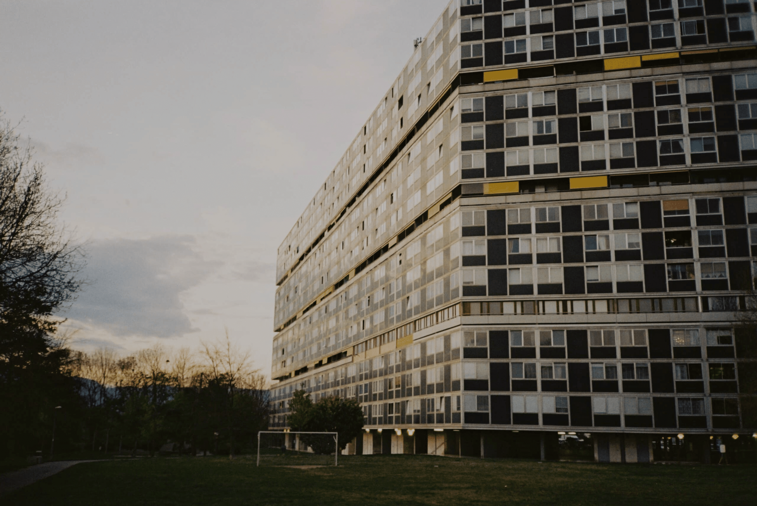 Grand immeuble résidentiel à nombreuses fenêtres, près d'un terrain de football et d'arbres au coucher du soleil.