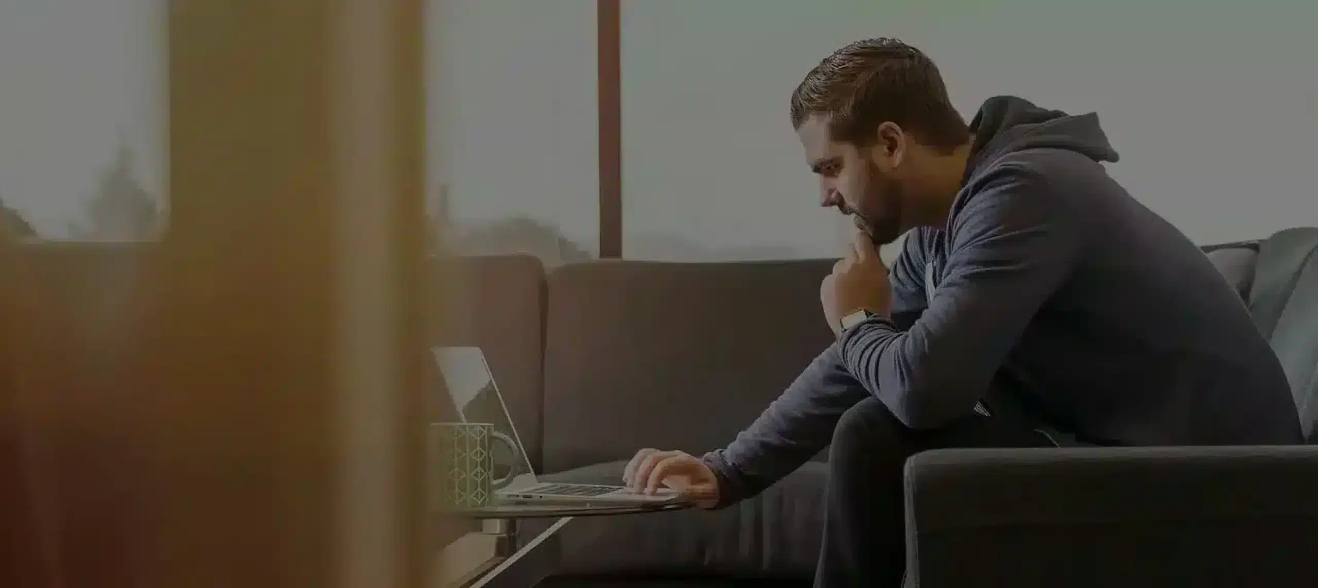 Homme assis sur un canapé utilisant un ordinateur portable sur une table basse avec une tasse – Dépannage Informatique Genève