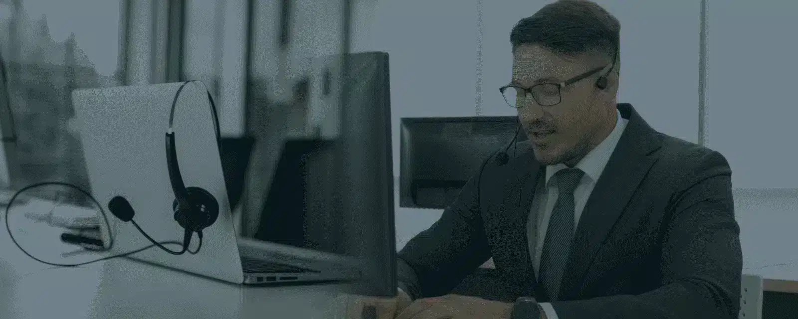 Homme en costume avec casque micro travaillant devant ordinateur et écran dans bureau moderne.